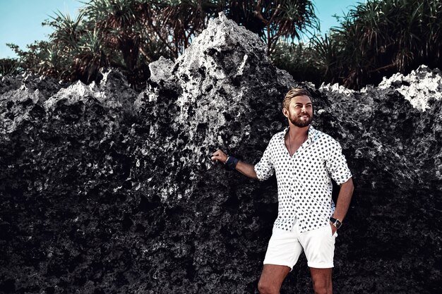 Portrait of handsome fashion man model wearing white clothes posing near rocks on the beach