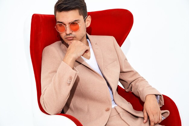 portrait of handsome fashion stylish businessman model dressed in elegant light pink suit sitting on red chair. Metrosexual