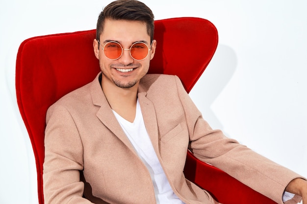 portrait of handsome fashion stylish businessman model dressed in elegant light pink suit sitting on red chair. Metrosexual