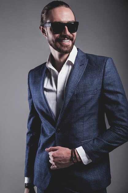 portrait of handsome fashion stylish hipster businessman model dressed in elegant blue suit in sunglasses posing on gray