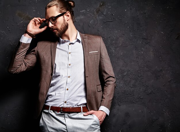 portrait of handsome fashion stylish hipster businessman model dressed in elegant brown suit in glasses near dark wall