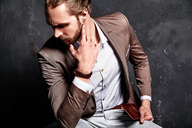 Портрет красивого модного стильного хипстерского бизнесмена, одетого в элегантный коричневый костюм, сидящего в темноте в студии