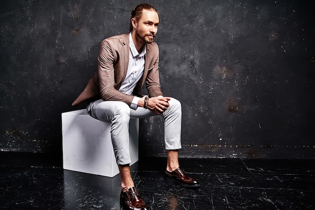 portrait of handsome fashion stylish hipster businessman model dressed in elegant brown suit sitting near dark in studio