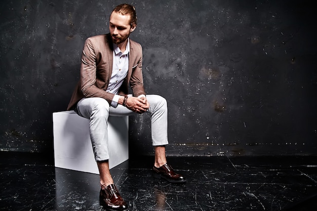 portrait of handsome fashion stylish hipster businessman model dressed in elegant brown suit sitting near dark in studio