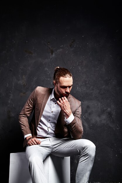 portrait of handsome fashion stylish hipster businessman model dressed in elegant brown suit sitting near dark