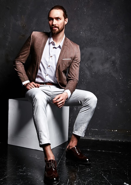 portrait of handsome fashion stylish hipster businessman model dressed in elegant brown suit sitting near dark