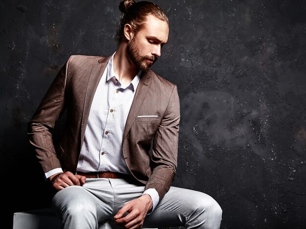 portrait of handsome fashion stylish hipster businessman model dressed in elegant brown suit sitting near dark