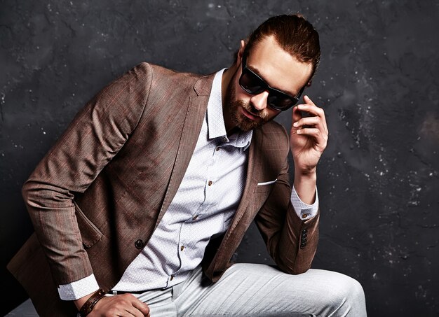portrait of handsome fashion stylish hipster businessman model dressed in elegant brown suit sitting near dark