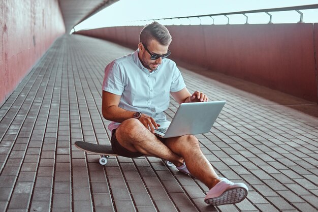 Portrait of a handsome fashionable freelancer in sunglasses dressed in a white shirt and shorts working on a laptop while sitting on a skateboard under a bridge.