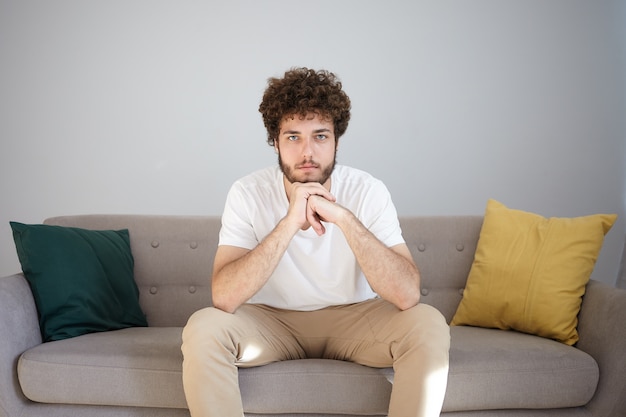 Free photo portrait of handsome fashionable young bearded guy in his twenties having rest indoors with calm facial expression, placing chin on clasped hands, sitting on comfortable couch