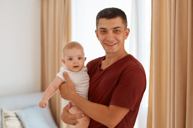 Free photo portrait of handsome father wearing burgundy t shirt with charming infant daughter in hands, looking at camera, standing in cozy living room with window on background.