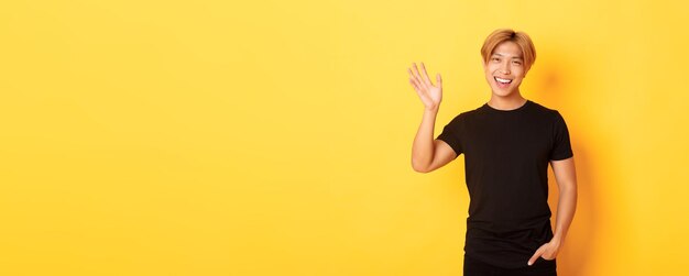 Free photo portrait of handsome friendly asian guy in black outfit waving hand to say hello and smiling greetin