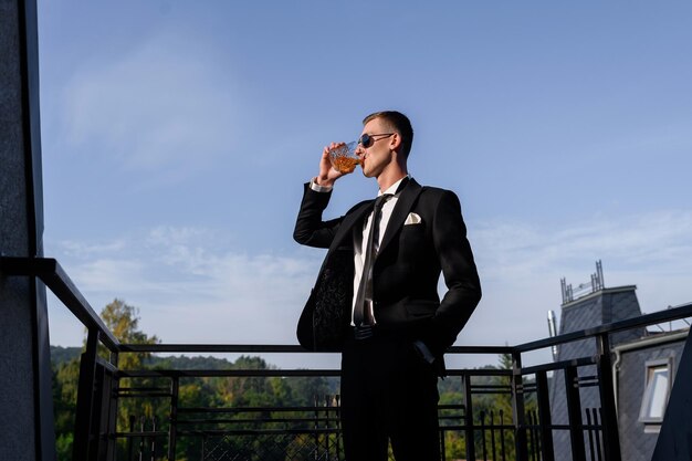Portrait of handsome groom man in stylish black suit standing on the balcony drinking looking at the sunset Celebrating wedding day Stag party Young attractive man in sunglasses Gentleman