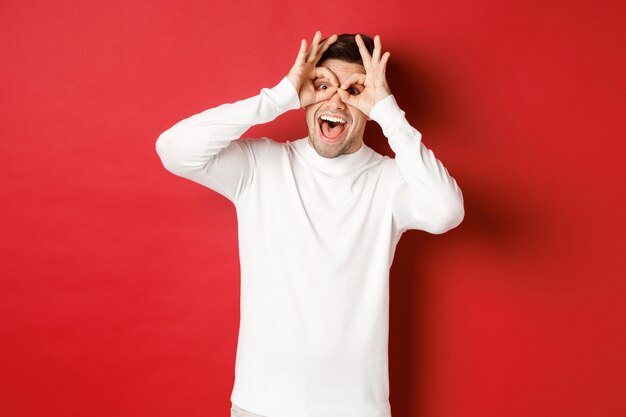 Portrait of handsome guy in white sweater making funny mask with fingers looking happy and smiling s...