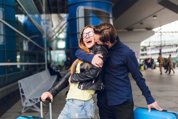 Portrait of handsome guy with beard in black shirt kissing girl with long hair outside in airport. She wears glasses, yellow sweater and jacket with jeans. She looks happy.