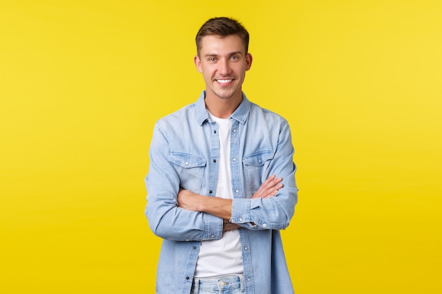 Portrait of handsome happy caucasian guy with white teeth, smiling broadly, cross arms chest confident, standing yellow background in denim shirt over white t-shirt.