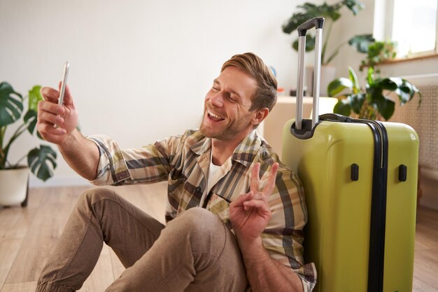 Portrait of handsome happy guy tourist takes selfies with suitcase uses smartphone