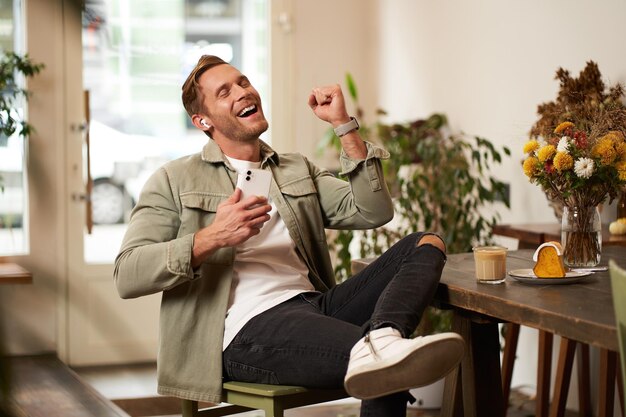 Free photo portrait of handsome happy man in cafe listens to music in wireless earphones holding smartphone