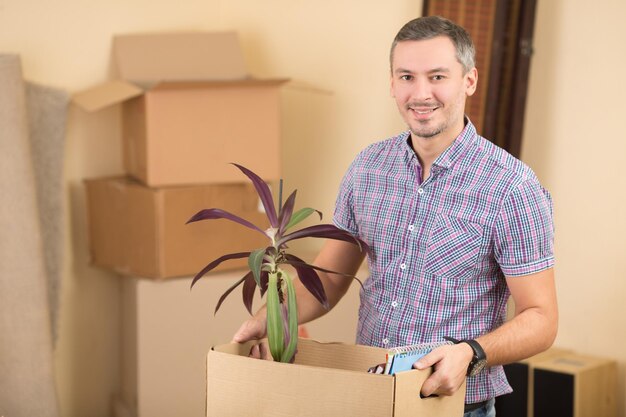 새 집으로 이사하는 동안 식물이 든 상자를 들고 있는 잘 생기고 행복한 남자의 초상화.
