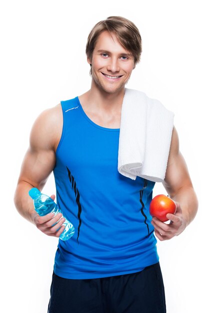 Portrait of handsome happy sportsman in blue shirt holds water and apple over white wall.