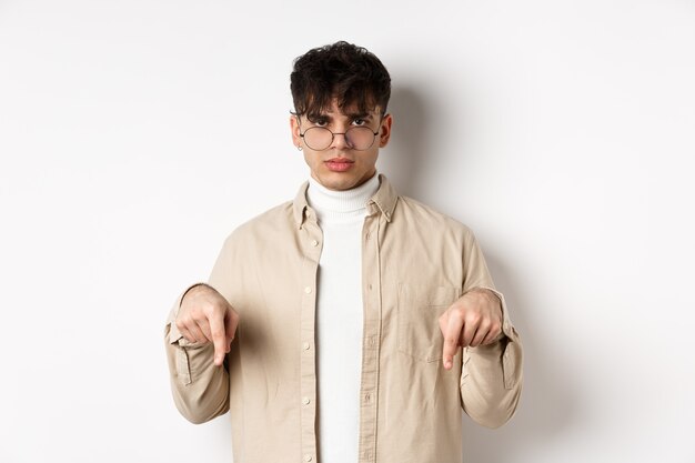 Portrait of handsome hipster guy in glasses showing an ad, pointing fingers down at logo, standing on white background.