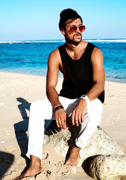 Portrait of handsome hipster sunbathed fashion man model wearing casual clothes in black T-shirt and sunglasses sitting on rocks