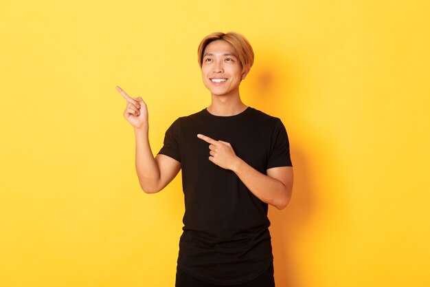 Portrait of handsome korean blond guy, pointing fingers upper left corner and looking satisfied with pleased smile, yellow wall.