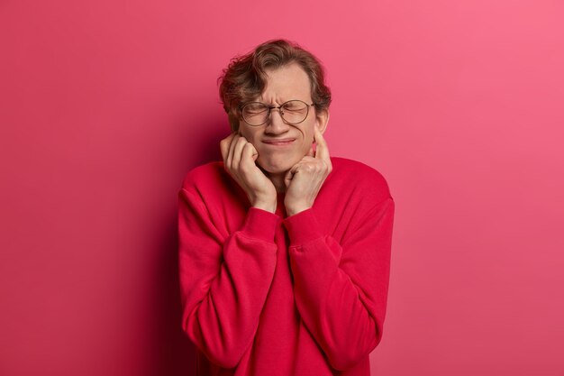 Portrait of handsome male youngster plugs ears, keeps eyes closed from displeasure, suffers from disgusting sound, ignores problems, smirks face, dressed casually, isolated on pink wall