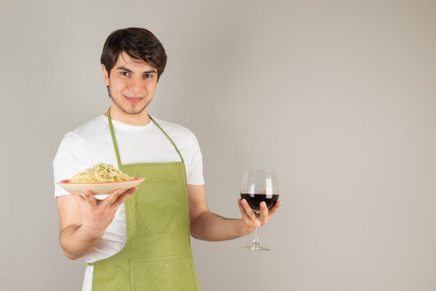 Portrait of a handsome man in apron holding a plate with noodles and a glass of wine.