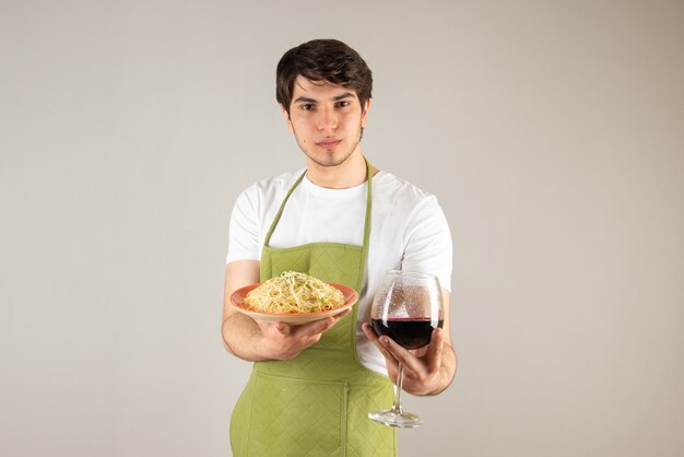 Портрет красивого мужчины в фартуке, держащего тарелку с лапшой и бокалом вина.