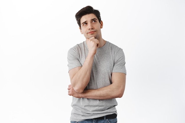 Portrait of handsome man in deep thoughts looking aside touching chin and thinking looking aside imaging something standing over white background