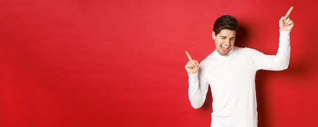 Portrait of handsome man enjoying new year party dancing and having fun pointing fingers up standing over red background