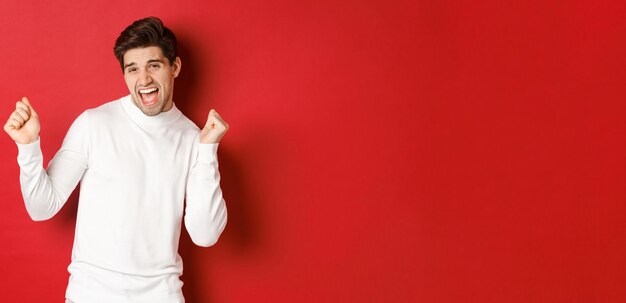 Portrait of handsome man enjoying new year party, dancing and having fun, standing in white sweater against red background