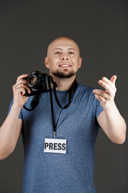 Portrait of handsome man in grey shirt with photocamera and press badge taking pictures over dark wall