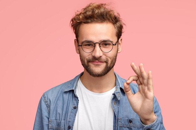 Portrait of handsome man has stubble, makes ok sign, agrees or likes something has joyful expression, poses against pink wall, proves everything goes according to plan. Body language concept
