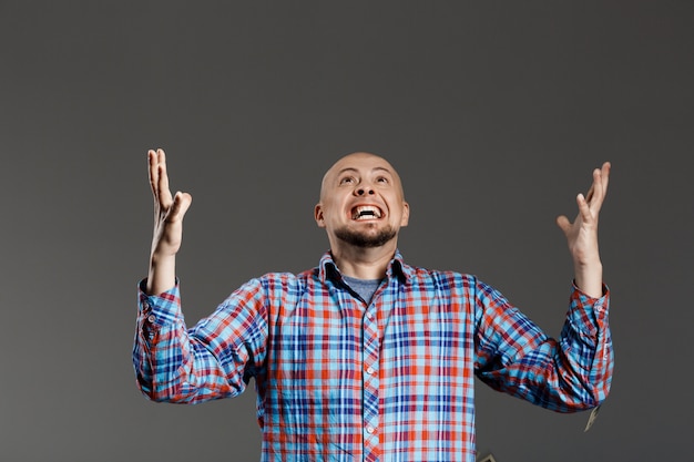 Portrait of handsome man screaming up lifting hands in plaid shirt over grey wall