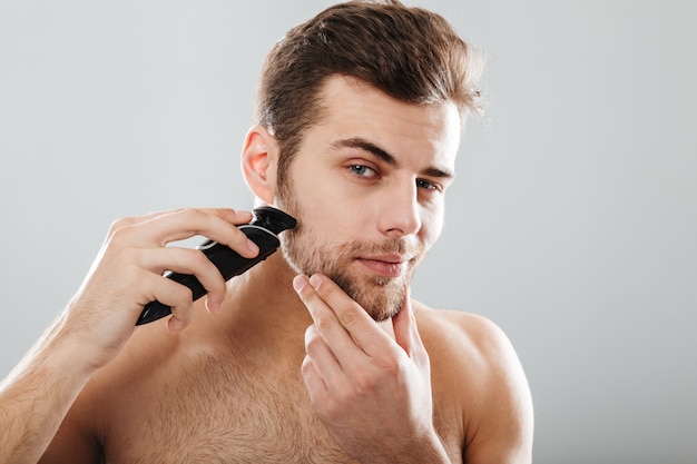 Foto gratuita ritratto dell'uomo bello che rade la sua barba con il rasoio elettrico nella mattina contro la parete grigia