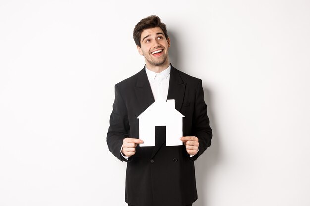 Портрет красивого мужчины в костюме, ищущего дома, держащего бумажный домик и мечтательного смотрящего на правый верхний угол, стоящего на белом фоне.