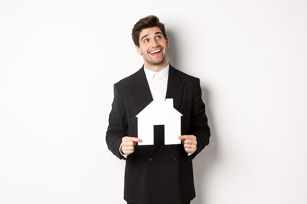 Portrait of handsome man in suit searching for home, holding paper house and looking at upper right corner dreamy, standing over white background.