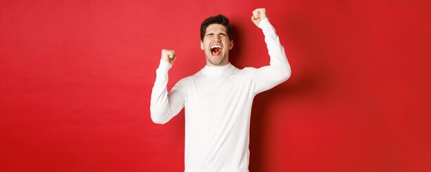 portrait of handsome man in white sweater, feeling cheerful, celebrating victory, shouting for joy and raising hands up in victory, standing over red background.