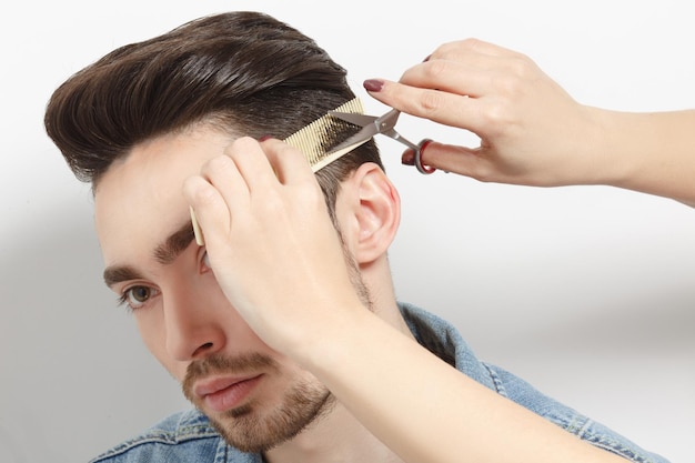 Portrait of handsome man with black hair having haircut in studio. Hairdresser using comb and scissors for creating modern hairstyle.