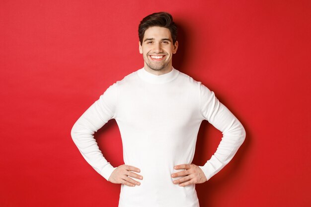 Portrait of handsome man with bristle, wearing white sweater, smiling and looking confident, standing against red background. Concept of new year and winter holidays.