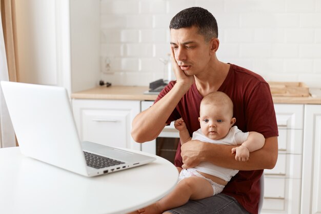 Portrait of handsome man with dark hair looking at notebook display with shocked face, wearing burgundy casual t shirt, working on laptop while babysitting, posing in white kitchen.