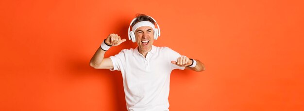 Portrait of handsome middleaged male athlete listening music in headphones during fitness training dancing over orange background