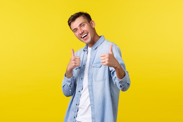 Portrait of handsome outgoing blond man showing thumbs-up in approval, encourage visit store. Male student inviting people to summer event or courses with special discount, yellow background.