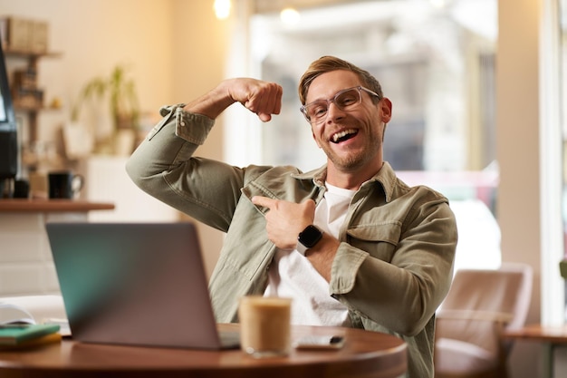 カフェのラップトップの前に座っているハンサムで満足した若者の肖像画バイセプスが筋肉を曲げている