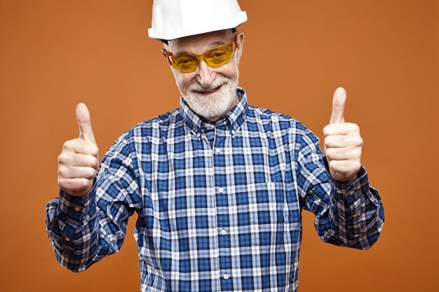 Portrait of handsome positive senior foreman in hardhat and yellow eyeglasses showing thumbs up gesture and smiling happily, encouraging his working crew for good job. Construction and renovation