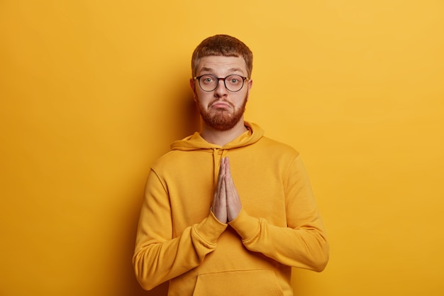 Portrait of handsome presses palms, makes praying gesture, pleads for help, purses lips and looks seriosly , dressed in casual hoodie, isolated over yellow wall. Please, do me favor