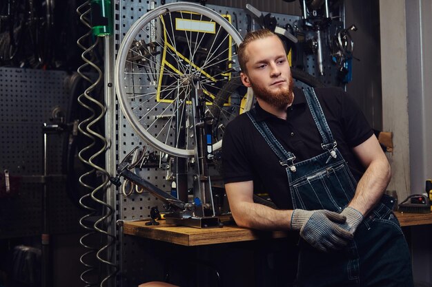 portrait of a handsome redhead male with beard and haircut wearing jeans coverall, standing near bicycle wheel in a workshop.