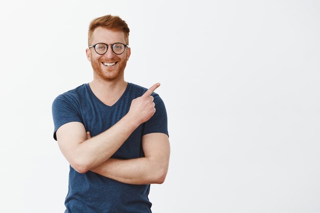 Portrait of handsome redhead male with bristle in glasses and t-shirt, pointing at upper right corner and smiling joyfully